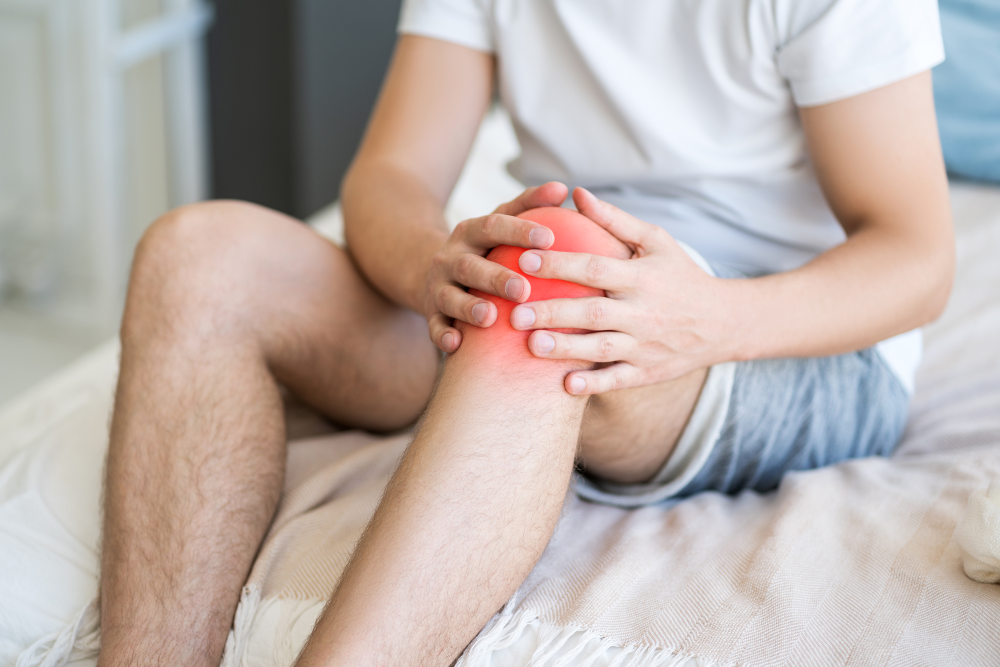 Person seated on a bed holding a knee, with soreness highlighted around the joint