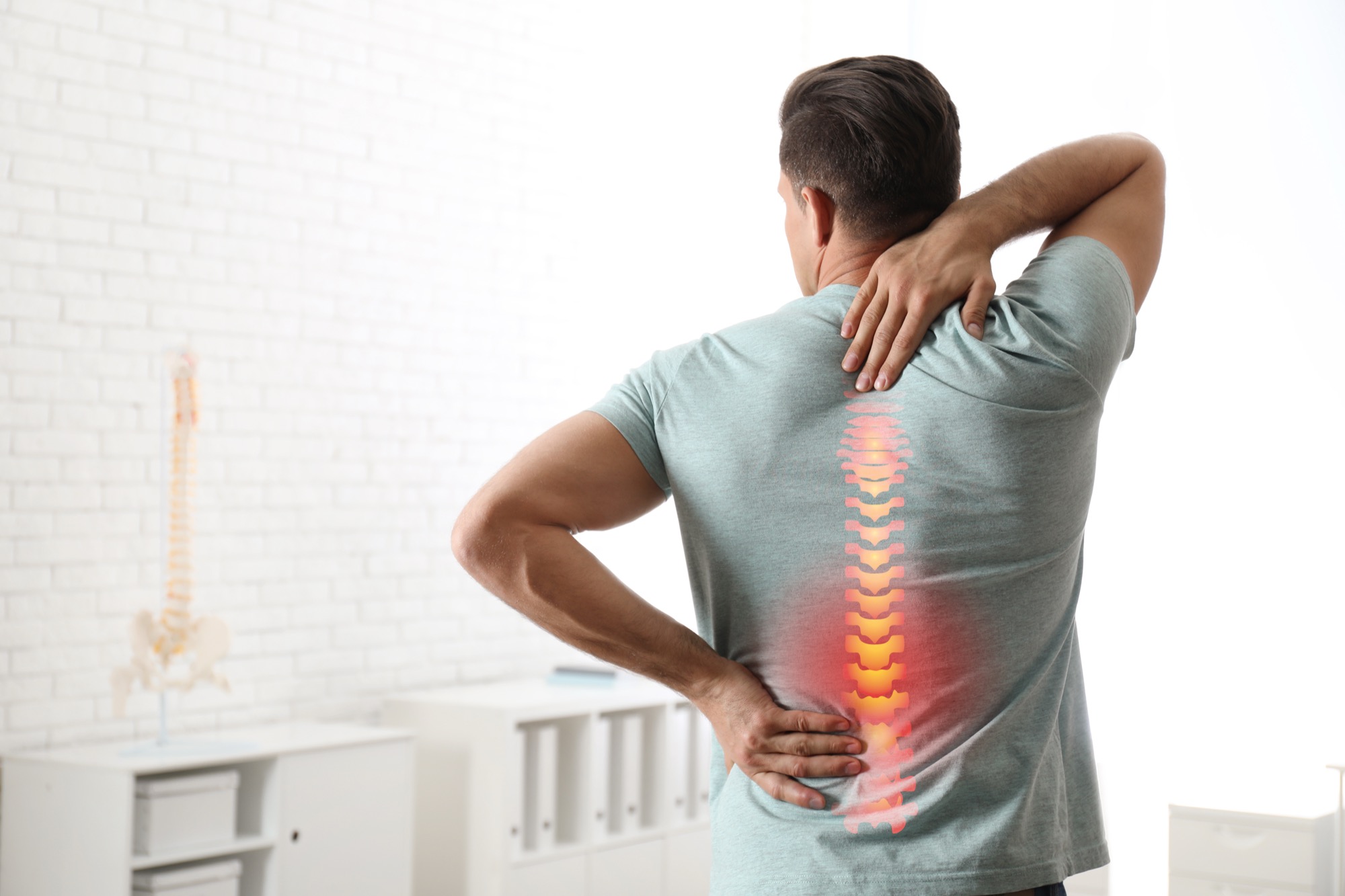 Man holding his neck and lower back, with the spine highlighted, in a clinic room with an anatomical spine model